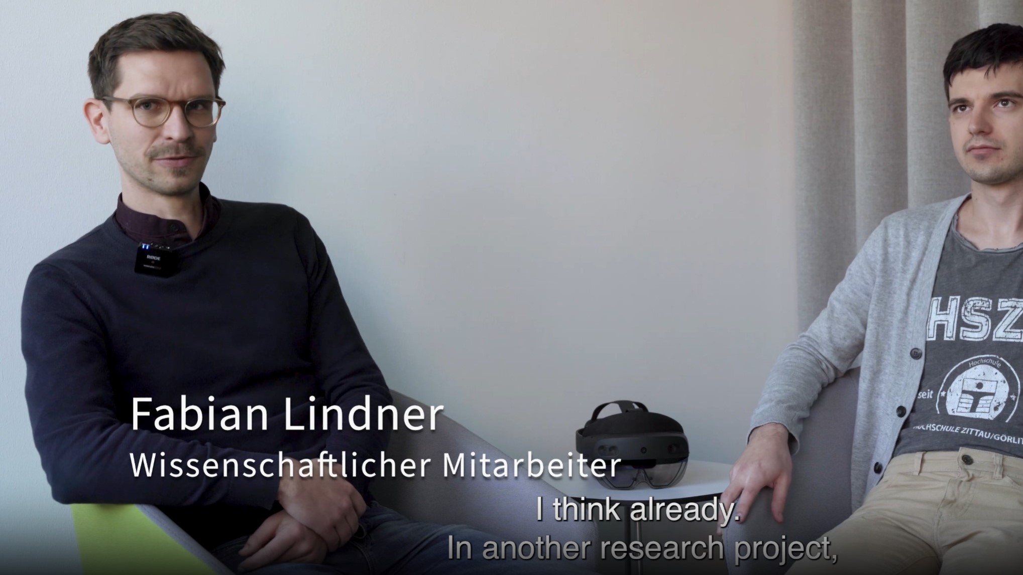 View the video |↗|. Photo: Infineon. A young man sits in a chair and looks into the camera while talking.